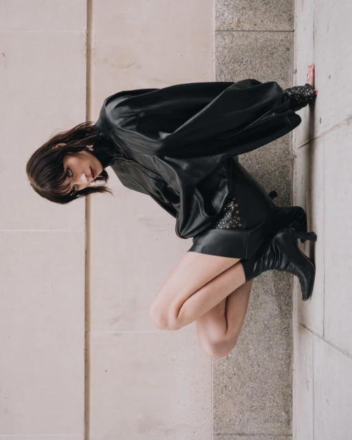 A fair-skinned woman with dark hair poses on a textured wall in a short black leather dress and boots, sideways in the fashion shot.