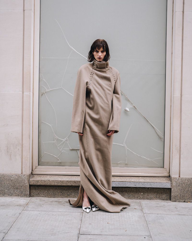 Fashion shot of a woman in a long beige coat in front of a shattered glass window on a city street, wearing shiny silver shoes.