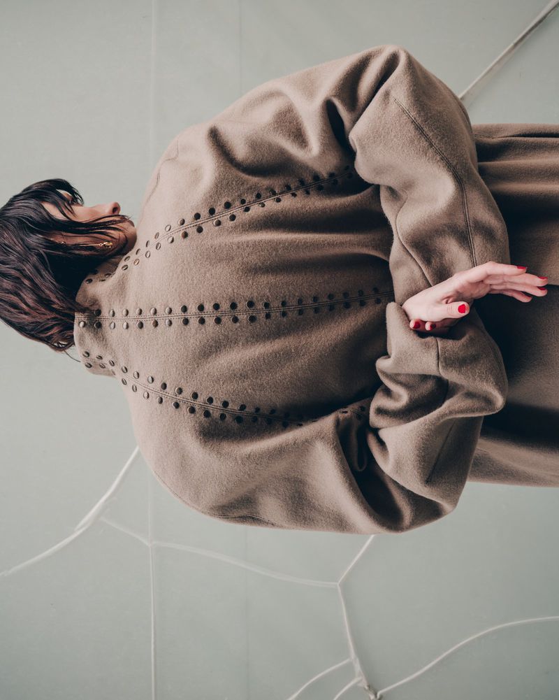 Brunette woman in a taupe coat poses on a light blue and white tile floor, showcasing a minimalist yet chic fashion aesthetic.