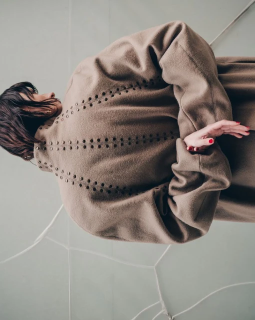 Brunette woman in a taupe coat poses on a light blue and white tile floor, showcasing a minimalist yet chic fashion aesthetic.