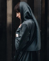 Profile of a woman wearing a dark hooded green garment and silver cuff bracelets against a dark wooden slatted background.