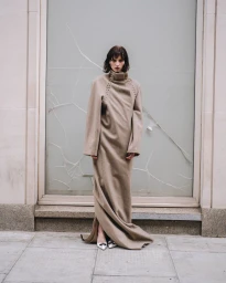 Model standing on stone paving wearing a long beige wool coat and pointed shoes against a cracked glass window.