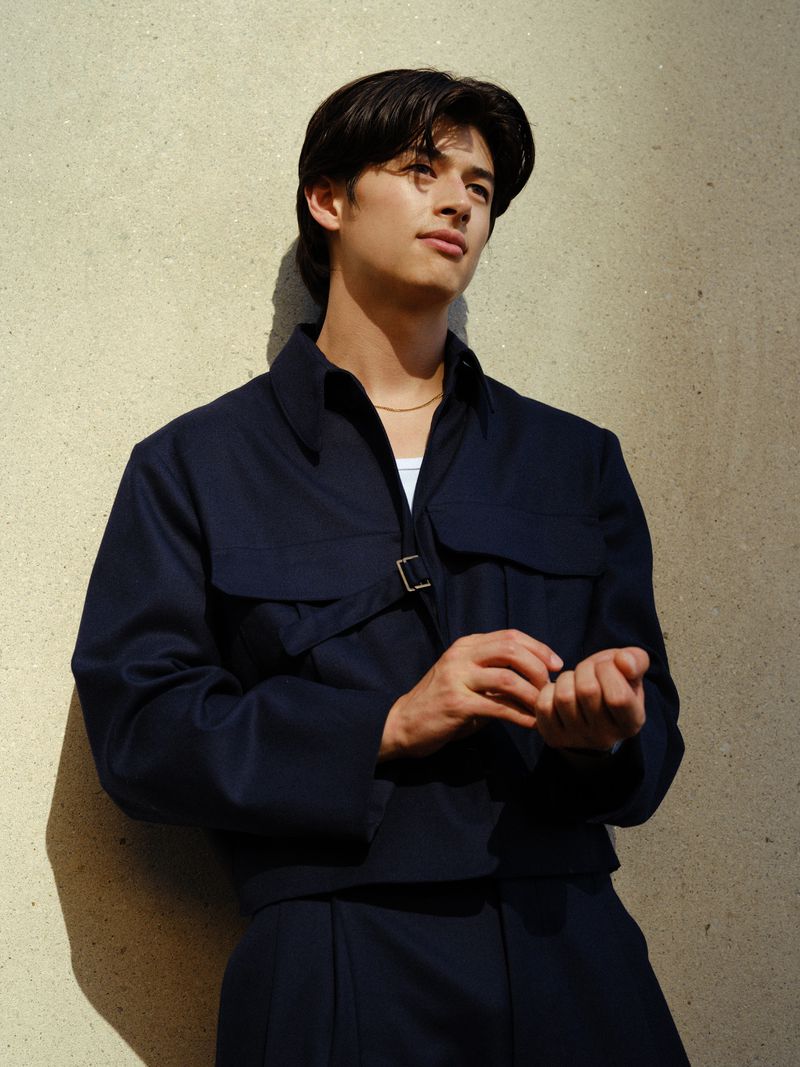 Portrait of a young adult male with dark hair wearing a navy blue jacket and pants, standing against a textured neutral wall with shadows.