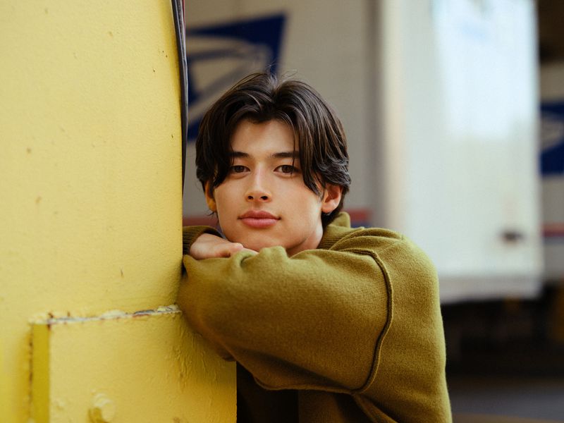 Portrait of an adult Asian man with dark hair resting his arms on a yellow pillar, looking at the camera with a neutral expression.