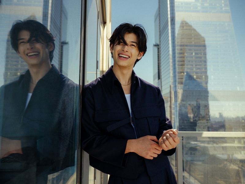 Smiling Asian man in a dark blue jacket poses outdoors with modern architecture in the background on a sunny day.