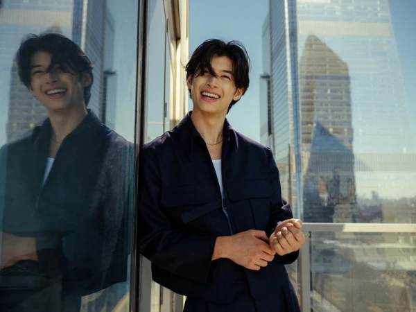 Smiling Asian man in a dark blue jacket poses outdoors with modern architecture in the background on a sunny day.