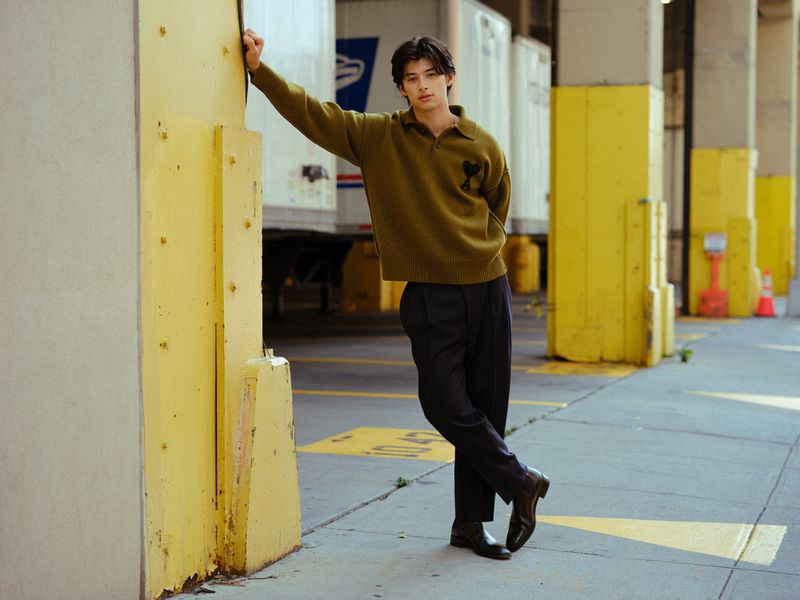 Stylish Asian man poses in an olive sweater and dress pants, leaning against yellow columns with semi-trucks in the background.