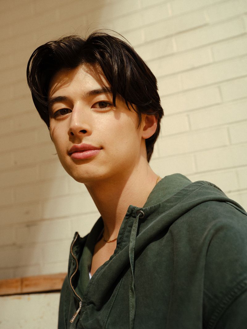 Portrait of a young adult male with dark hair wearing a green hooded zip-up jacket, looking at the camera, against a brick wall.
