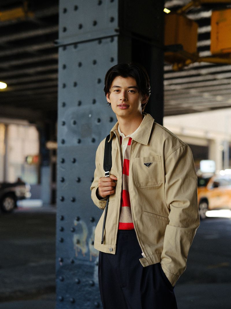 A young adult male stands confidently in an urban setting, wearing a tan jacket, striped shirt, and carrying a backpack, posing near a large metal column.