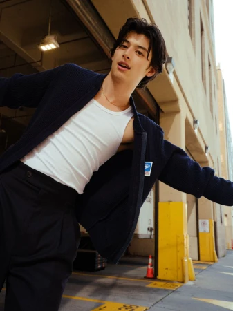 Young Asian male model in a white tank top and navy jacket poses in a sunny urban setting with yellow painted lines.
