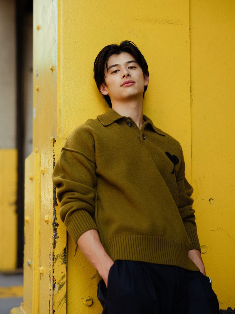 Portrait of a young adult male in a green collared sweater leaning against a yellow painted beam in an urban environment.