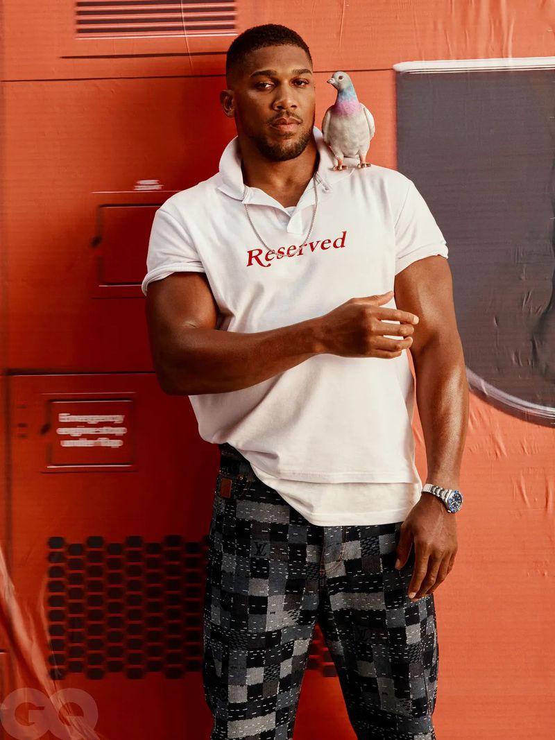 Anthony Joshua portrait with a grey pigeon perched on his shoulder, wearing a white polo shirt, dark pants, against a reddish-orange backdrop.