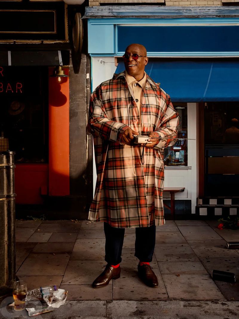 Bald adult male in a plaid poncho holds a small box in front of a building with a blue awning on a city street with trash on the ground.