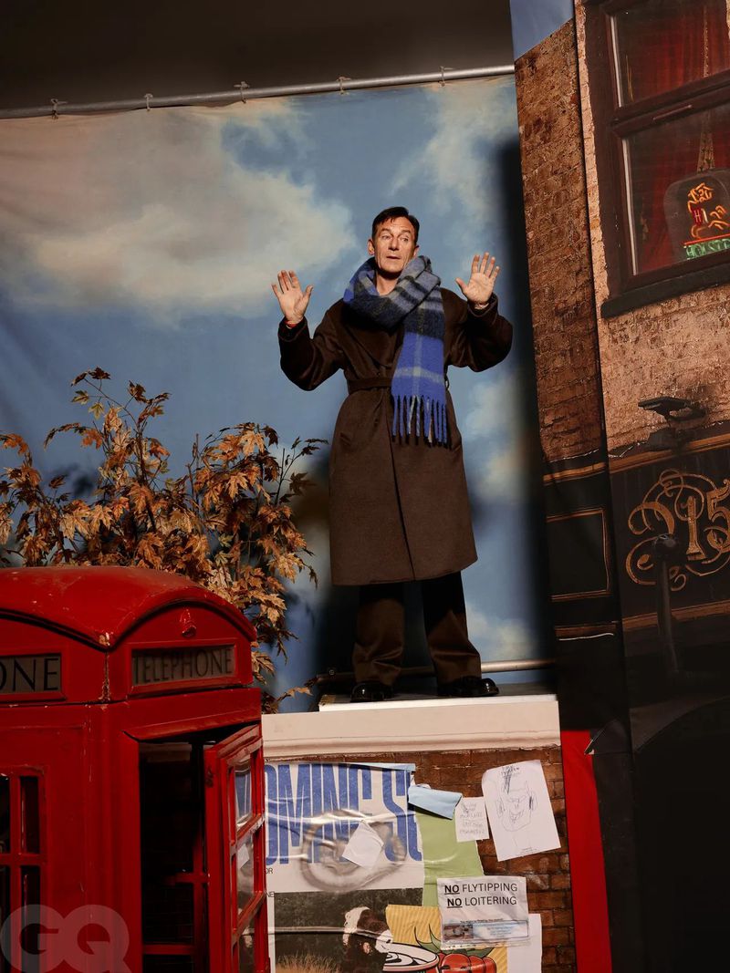 Adult man stands in brown robe and blue scarf in front of a painted studio backdrop with a red telephone booth prop.