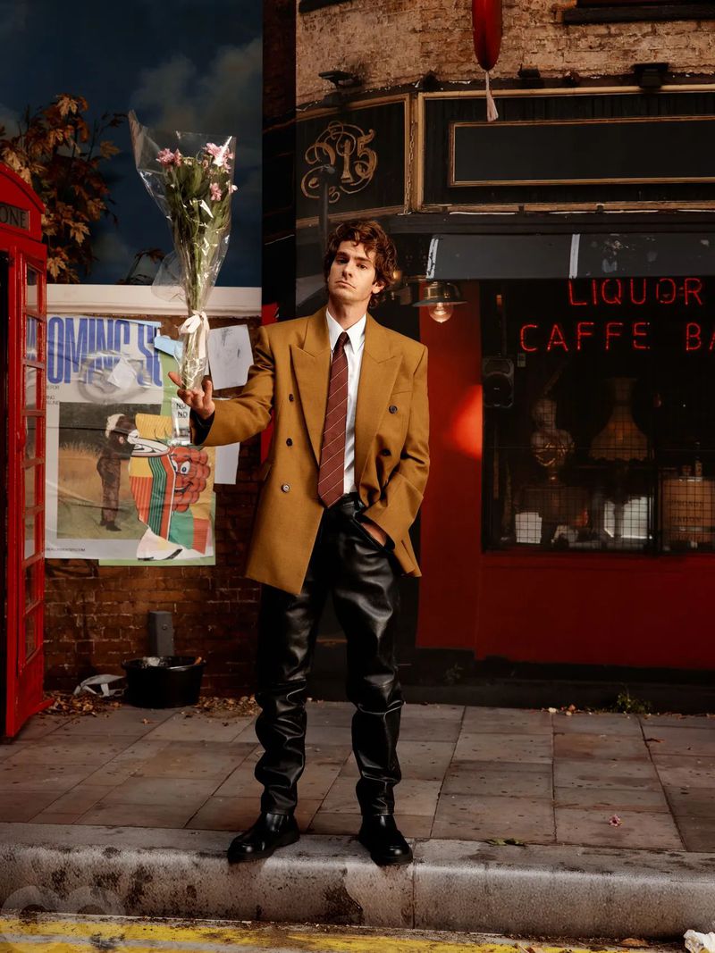 An adult man with curly hair in a double-breasted coat and leather pants standing in front of a storefront holding white flowers.