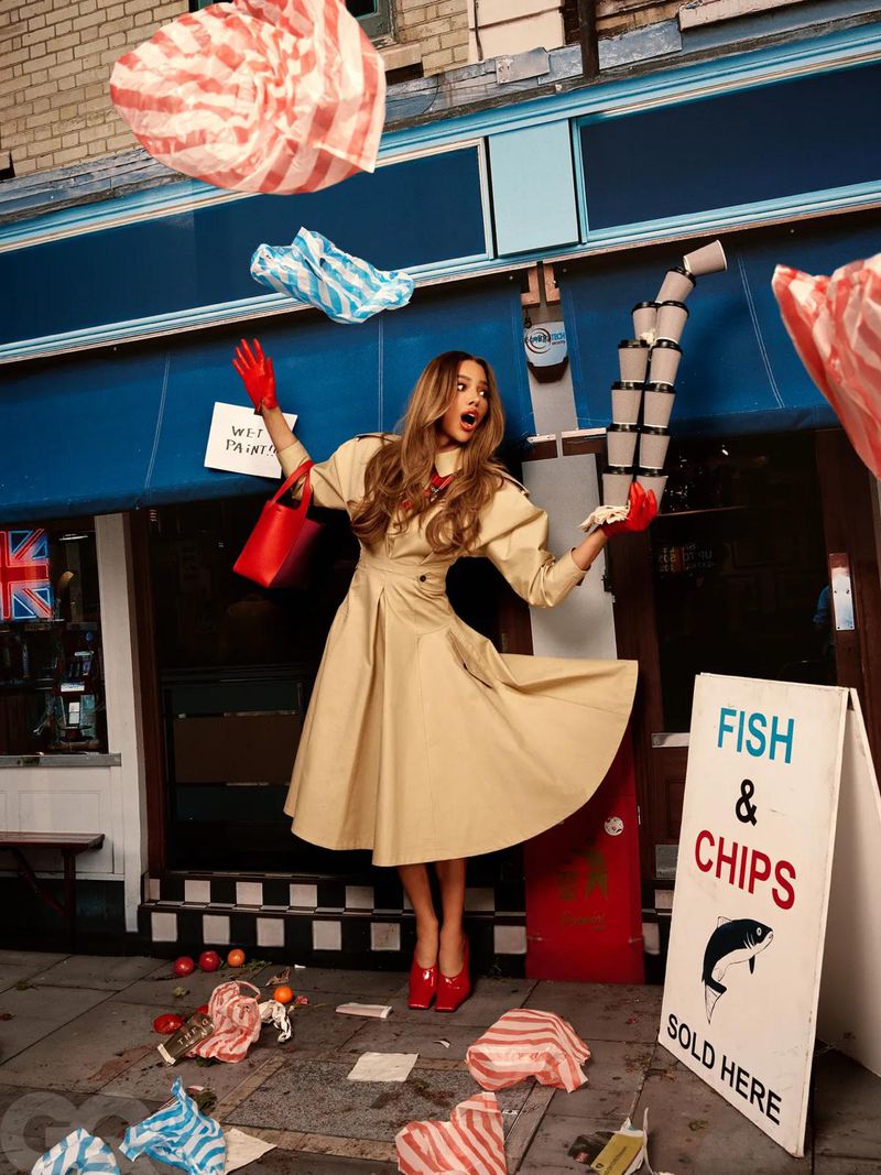 Stylish woman posing outside fish and chips shop with floating paper, wearing a beige dress, red gloves, and carrying a red purse.