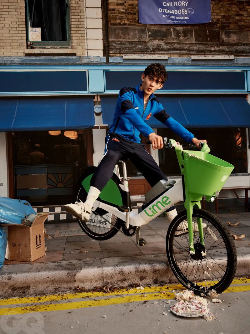 Young man on a Lime bike in an urban setting, wearing a blue tracksuit, captured in a medium shot on a city street with brick buildings.