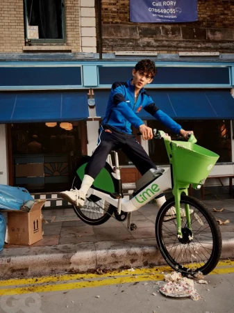 Young man on a Lime bike in an urban setting, wearing a blue tracksuit, captured in a medium shot on a city street with brick buildings.