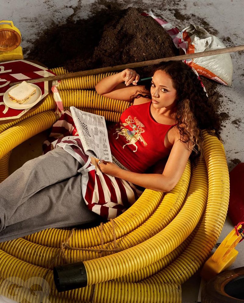 Woman lounges amidst yellow flexible tubes in an eclectic setting. She reads a newspaper crossword puzzle with construction items nearby.