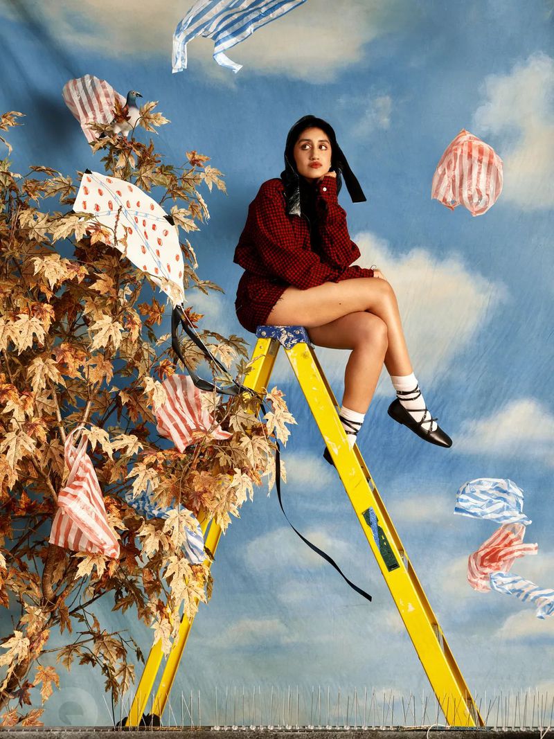 Young woman in red sweater and black flats sits on a yellow ladder next to a tree with small patterned fabrics in front of sky backdrop.