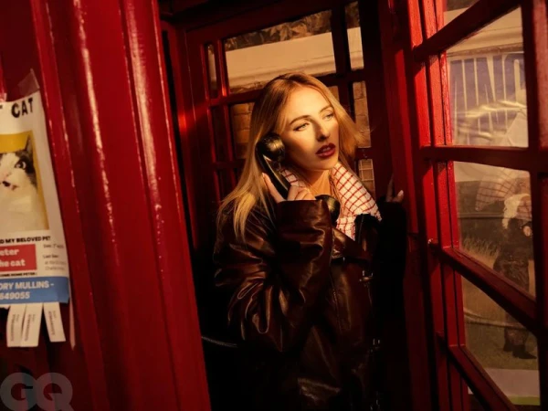 Blonde woman in a brown leather jacket using a vintage rotary phone in a red telephone booth with a lost cat poster.