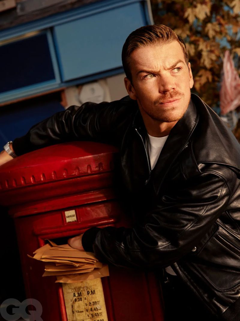 Portrait of a man in a leather jacket leaning on a red British postbox outdoors with golden light and fall foliage in the background.