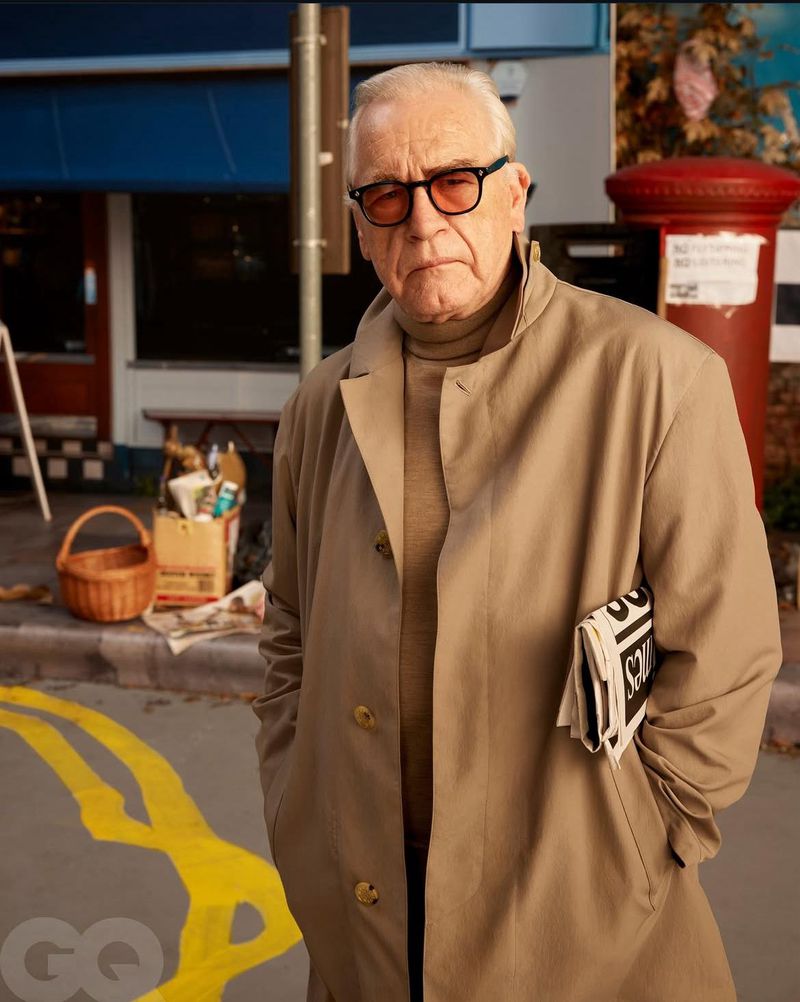 Portrait of a mature man wearing a trench coat and eyeglasses, holding a newspaper in an urban setting with street details.