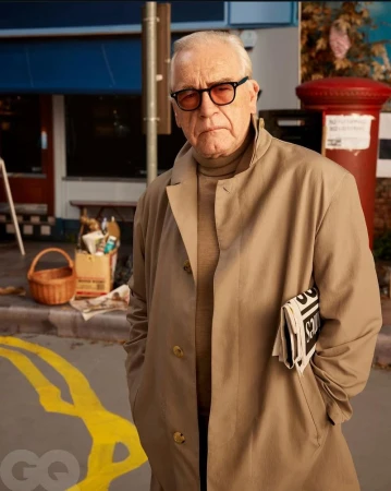 Portrait of a mature man wearing a trench coat and eyeglasses, holding a newspaper in an urban setting with street details.