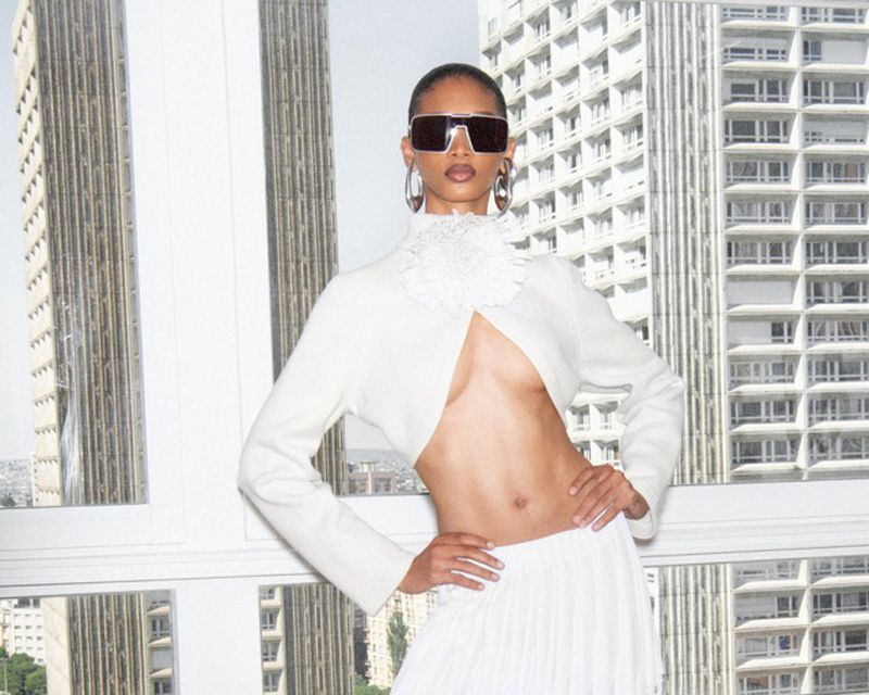 Woman posing in white bolero jacket and pleated skirt against a city background, wearing sunglasses and hoop earrings.
