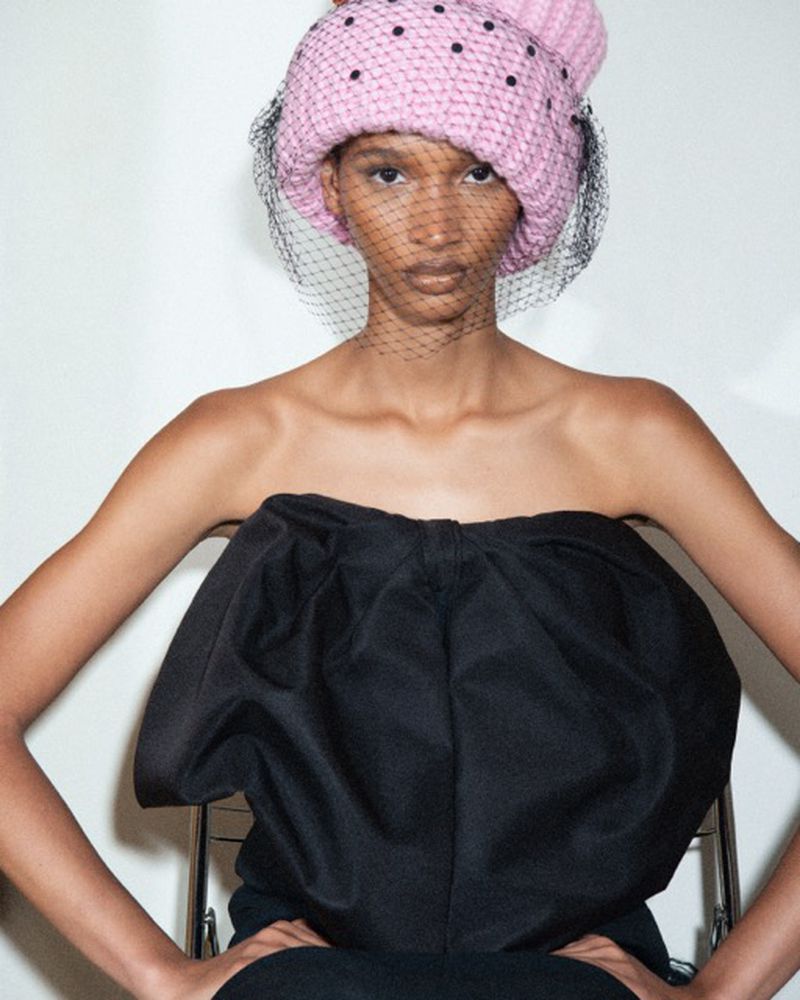 A fashion shot of a slender woman with a polka-dot hat and netting, wearing a strapless black top, seated indoors against a white wall.