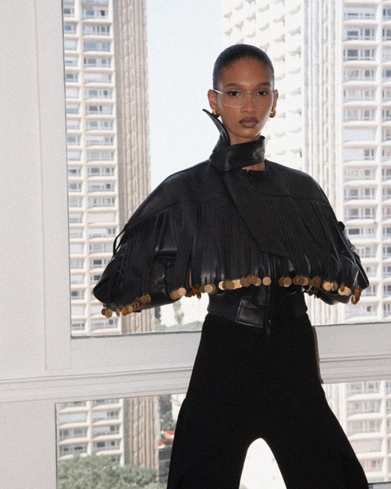 An adult female model poses in an open black leather jacket with gold coin detailing and black pants in front of a city view window.