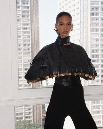 Model wearing a black leather fringed jacket against a large window overlooking urban high-rise buildings.