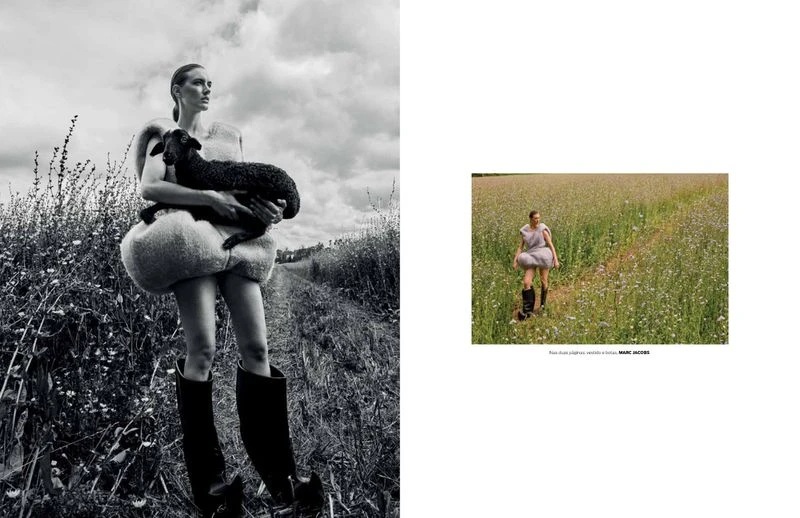 A woman in a dress and boots holds a dark lamb in a black-and-white outdoor editorial fashion shot with tall grass and cloudy sky.
