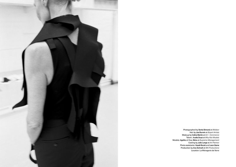 Black-and-white fashion shot of a person wearing a dark sleeveless vest with a sculptural flap detail and black pants in an indoor setting.