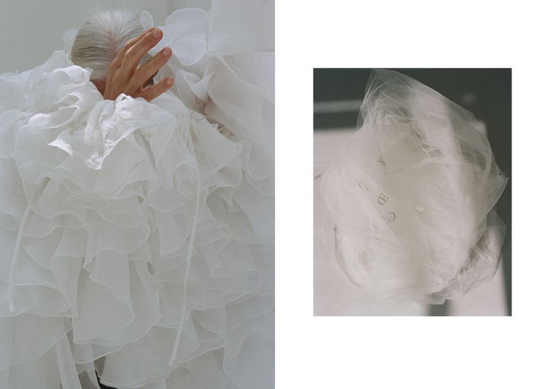 Silver-haired woman wearing a white, ruffled garment holds her hand up in front of her face, posing in a photo studio setting.