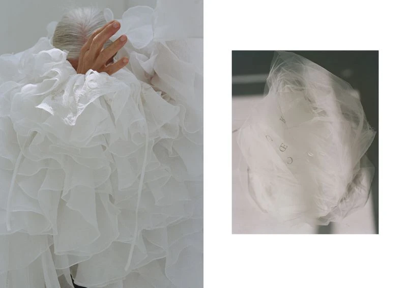 Silver-haired woman wearing a white, ruffled garment holds her hand up in front of her face, posing in a photo studio setting.
