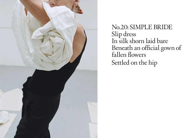Adult woman wearing a black sleeveless top and pants with a large, folded, flower-like white garment over her shoulders in a minimalist setting.