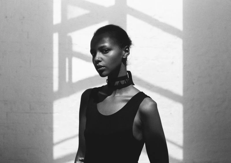 Portrait of a Black woman with geometric shadows on a white wall, wearing a choker and a black top in a black and white photo.