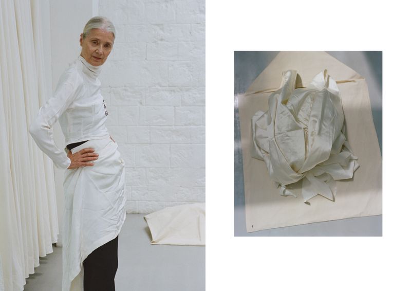 Older woman models white long-sleeve top and draped skirt indoors in front of white brick wall next to a white fabric sample.