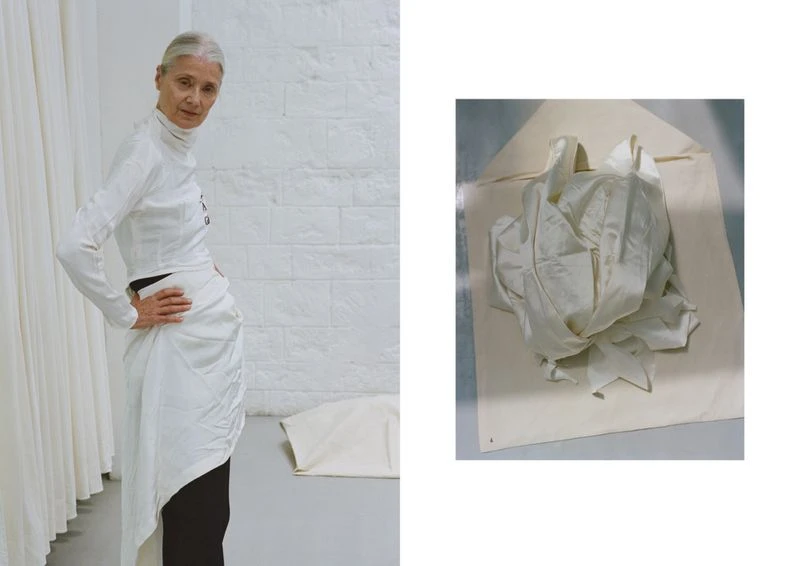 Older woman models white long-sleeve top and draped skirt indoors in front of white brick wall next to a white fabric sample.