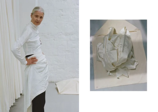 Older woman models white long-sleeve top and draped skirt indoors in front of white brick wall next to a white fabric sample.