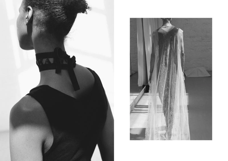 Black and white fashion shot of an adult woman wearing a black choker and a woman in a sheer dress standing indoors.