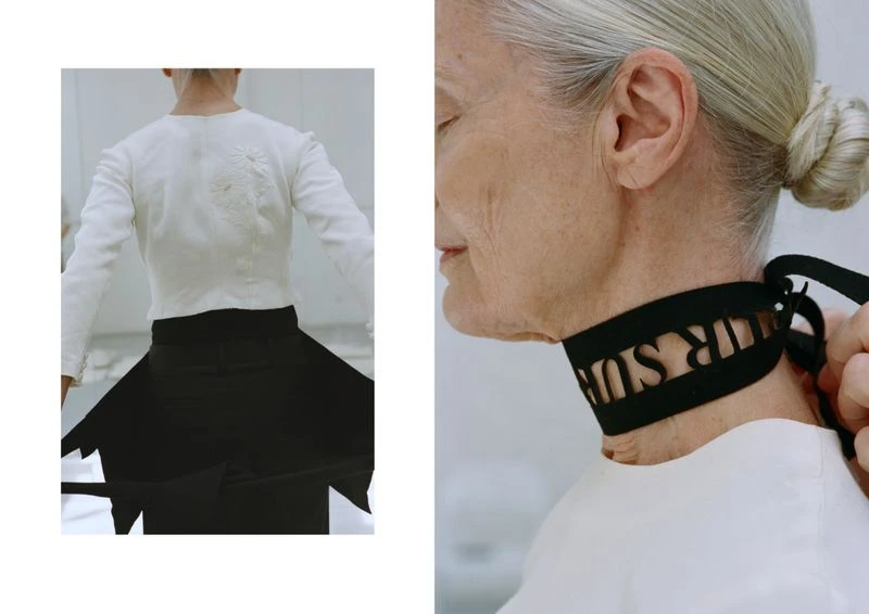 Elderly woman models a couture black ribbon choker with cutout letters, wearing a white top against a minimalist white backdrop.