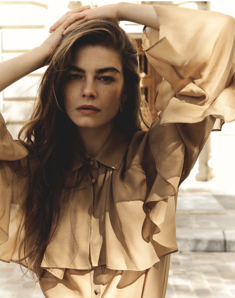 Elegant woman with freckles poses hands behind her head, wearing a tan ruffled button-down blouse in soft light against an outdoor background.