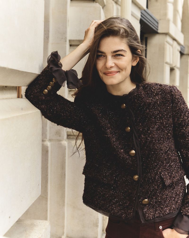 Smiling woman with long brown hair poses in a dark tweed jacket with gold buttons against a light stone wall in an outdoor setting.