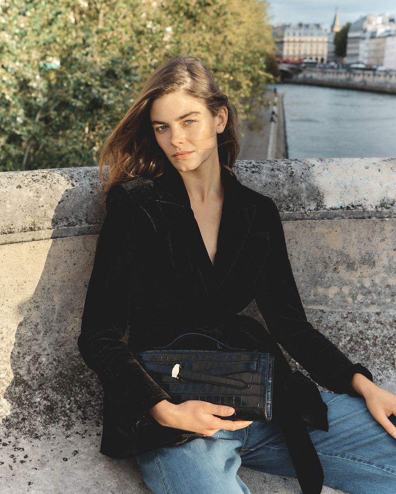 A woman in a black blazer sits on a stone wall, holding a black clutch bag with a city view in the background, fashionably styled.