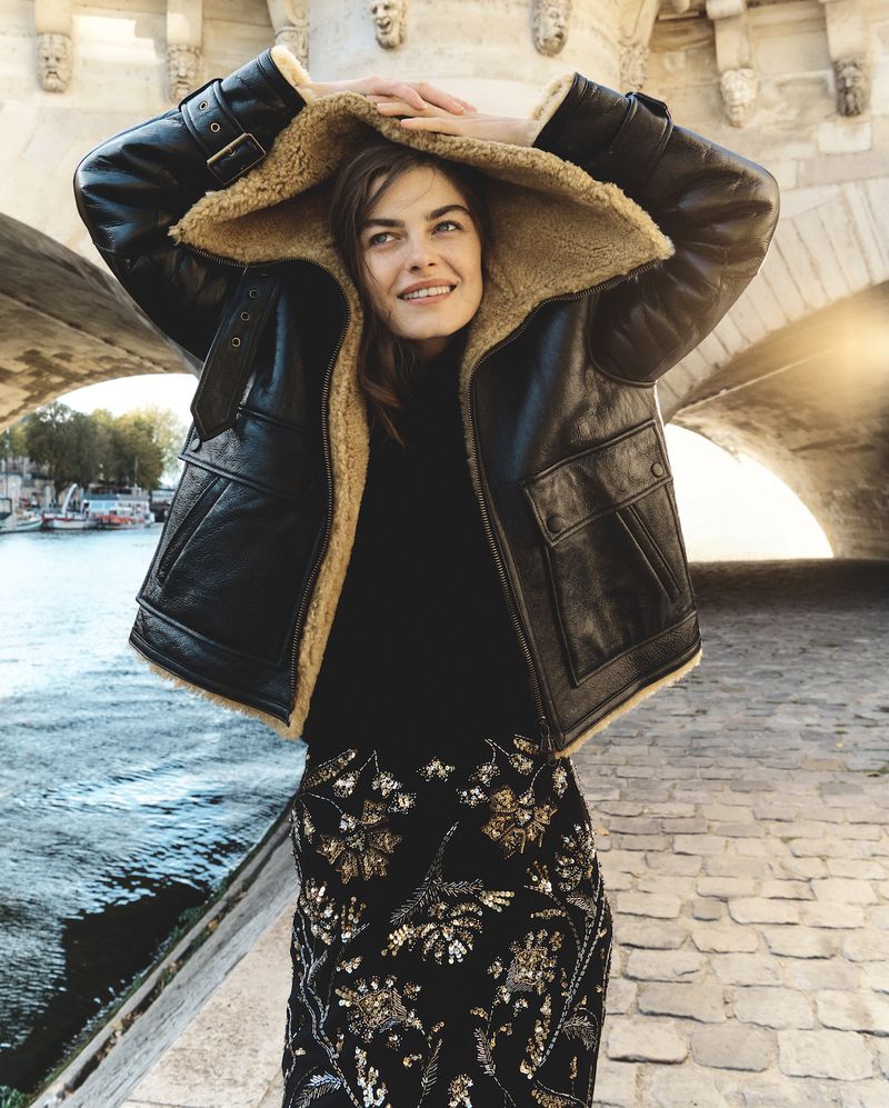 Model wearing a black shearling jacket with her hood up, standing by a stone bridge over a river.