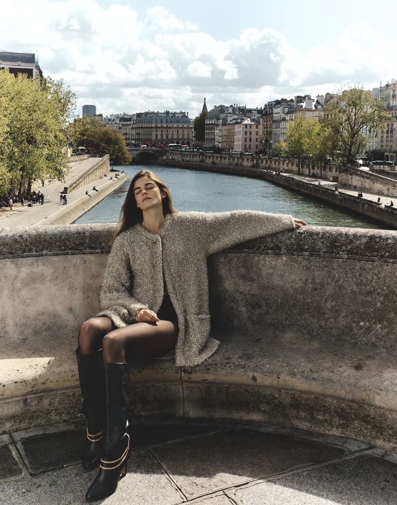 A woman in a coat and boots poses on a bridge over a river in Paris, with historic buildings and a blue sky in the background.