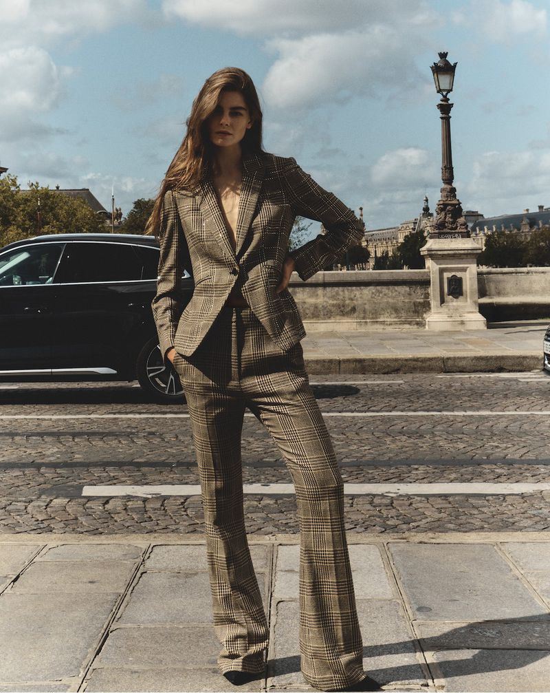 A brunette woman poses in a plaid suit on a bridge in Paris, with a street, black car, and architectural details in the background under a cloudy sky.