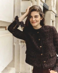 Model leaning against a pale stone building wearing a dark tweed jacket with metallic buttons for Veronica Beard Fall 2025.